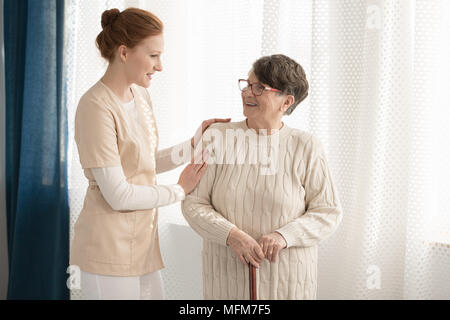 Professional caregiver in uniform talking to and assisting an elderly woman with a cane standing against white background in a retirement home Stock Photo