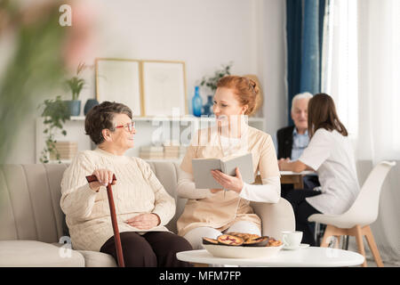 Professional caretaker in uniform reading a book to a happy senior woman with cane while sitting on a couch during leisure time in luxury nursing home Stock Photo