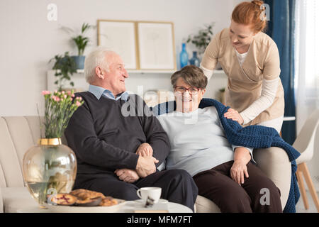Cheerful senior couple sitting on a couch with a tender caregiver standing next to them and putting a blue blanket over the woman's shoulder in a brig Stock Photo