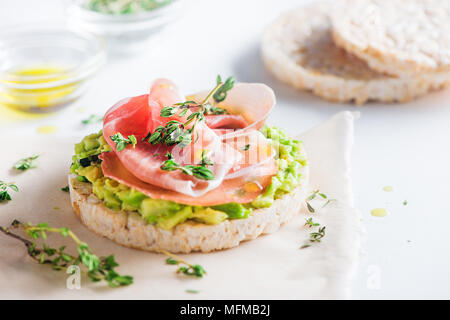 Crisp bread healthy snack with Parma ham, avocado spread, olive oil, thyme. Easy breakfast close-up on a white background with copy space. Stock Photo