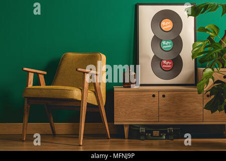 Retro armchair standing next to a wooden cabinet with a poster on top set on the dark green wall Stock Photo