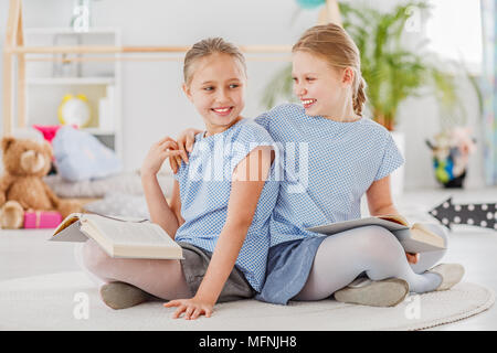 Girl laughing with an arm around her sister's shoulder, sitting on a white rug and reading together Stock Photo