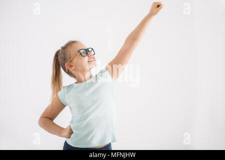 Portrait of a young girl with her arm and fist up and the other arm on a hip in a superhero pose on white wall background Stock Photo
