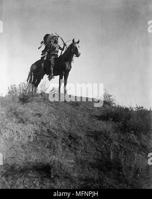 Native American Indian man in feather headdress, on horseback, holding bow and arrows, 1 arrow in his  mouth, 1908.  Photograph by Edward Curtis (1868-1952). Stock Photo