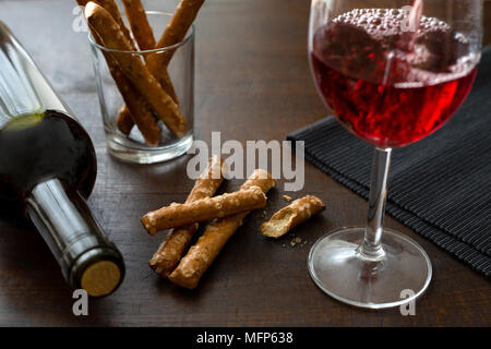 Pouring red wine into a glass next to pretzel sticks and wine bottle on rustic dark wooden table. Stock Photo