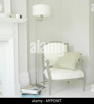Detail of a traditional style grey wood framed armchair with white upholstery, next to a tall lamp and a fireplace in a sitting room with white walls Stock Photo