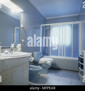 Bathroom with a tiled floor and a blue toilet and bidet in front of a bath with a shower screen. Stock Photo