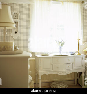 Traditional bedroom detail white period furniture including a small dresser with a vase of lavender on top in front of a window. Stock Photo