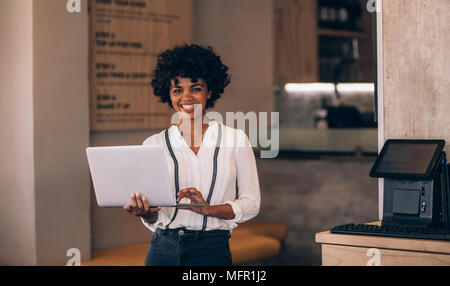 Portrait of young african woman with a laptop standing in her restaurant. Cafe owner using laptop and smiling. Stock Photo