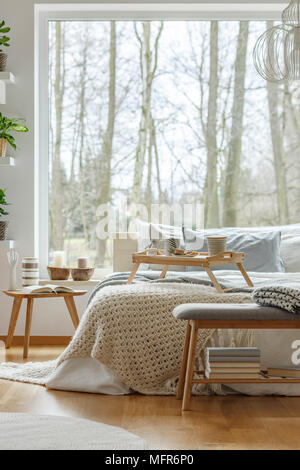 Knit blanket on bed next to wooden table against the window in natural bedroom interior of a house Stock Photo