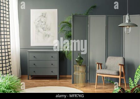 Dark, gray living room interior with a sketch hanging above a wooden chest of drawers next to a screen, golden table and armchair Stock Photo