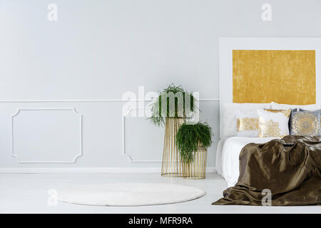 Potted plants on modern design, golden metal tables next to a cozy double bed in a minimalist bedroom interior with abstract painting Stock Photo