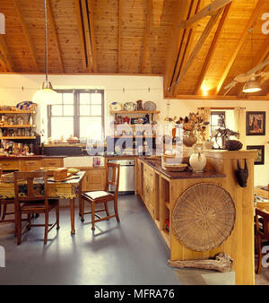 Split level open plan country kitchen in barn conversion neutral wall sloping wood ceiling units shelves table chairs white Belfast sink kitchenware l Stock Photo