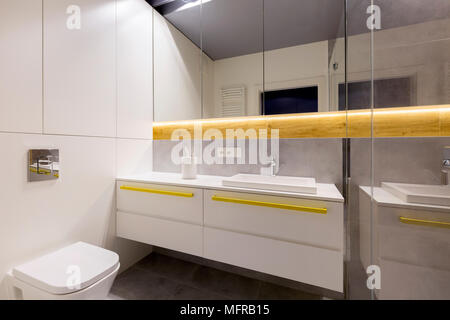 Clean, modern bathroom interior with a toilet, white cabinets, yellow handles, mirrors and gray tiles Stock Photo