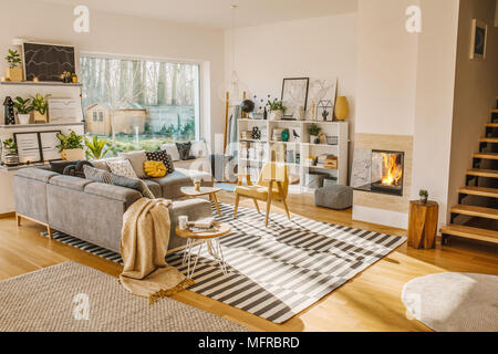 Blanket thrown on grey corner sofa in white living room interior with fresh plants, carpet, fireplace and simple posters Stock Photo