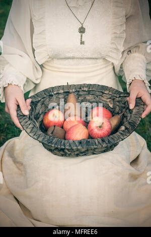 a woman in a white edwardian dress is sitting on a meadow and holding a basket with fruits on her lap Stock Photo