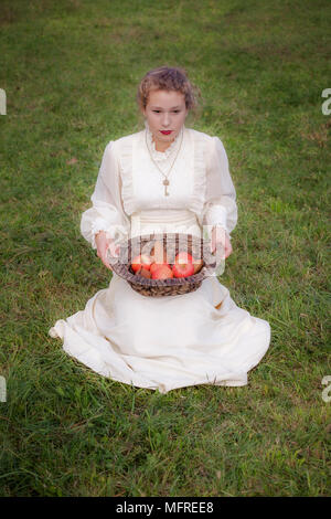 a woman in a white edwardian dress is sitting on a meadow and holding a basket with fruits on her lap Stock Photo