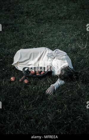 a woman in a white edwardian dress is lying in the grass, in front of her a basket with fruits Stock Photo