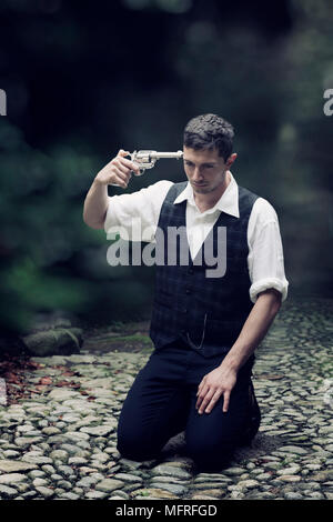 a man in vintage clothes is sitting on the ground, trying to shoot himself Stock Photo