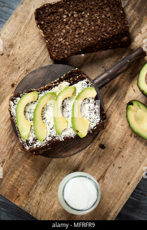 high angle view of a slice of rye bread topped with some slices of avocado, and a glass jar of yoghurt, on a rustic wooden table Stock Photo