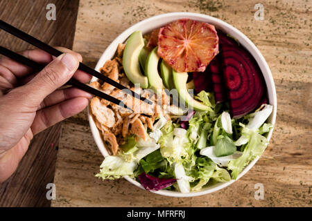 closeup of a young caucasian man picking some chicken with chopsticks from an appetizing buddha bowl, placed on a wooden table Stock Photo