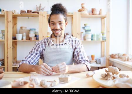 Portrait of young handsome mixed race craftsman smiling at camera while sitting at table and shaping clay in workshop Stock Photo