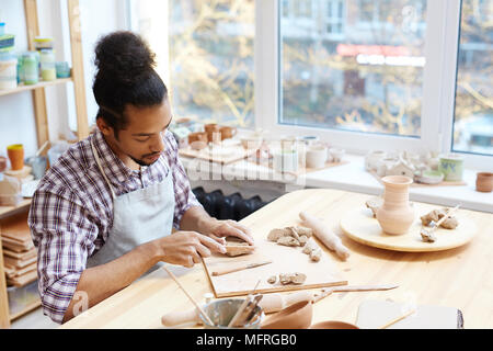 Young concentrated mixed-race male ceramist preparing clay while modeling pots in workshop Stock Photo