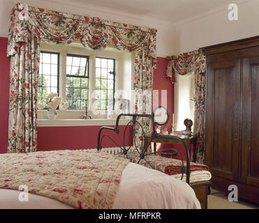 Country bedroom with floral swag curtains, wrought iron bed, and a dark wood wardrobe. Stock Photo