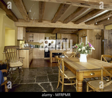 Country, yellow kitchen and dining area with beamed ceiling, wooden cabinets, dining table and chairs, and a flagstone floor. Stock Photo