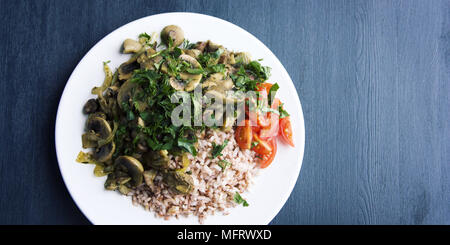 Red rice with mushrooms and cherry tomatoes. Vegan dish. European cuisine. Simple healthy lunch on the black background. Vegetarian rice dish on the w Stock Photo