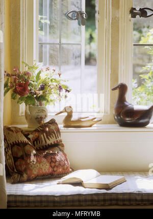 Open book and cushion on window seat with ornamental ducks and flower arrangement on ledge. Stock Photo