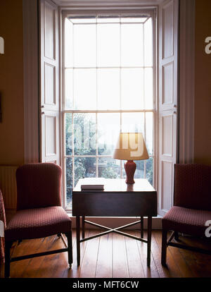 Traditional sitting room with a wooden side table and upholstered chairs in front of a sunny bay window with open shutters. Stock Photo