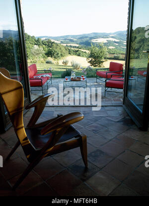 Tiled sunroom with a wooden armchair and open glass doors to a patio with a view. Stock Photo