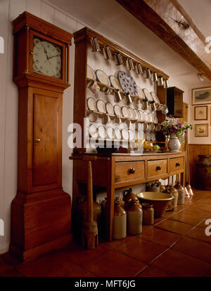 Country kitchen with tile floor, wooden buffet with open display shelves, collection of china and crockery, and an antique grandfather clock. Stock Photo