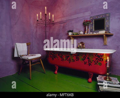 Dramatic purple bathroom with freestanding roll top bathtub, wooden chair, lit candelabra, and green painted floorboards. Stock Photo