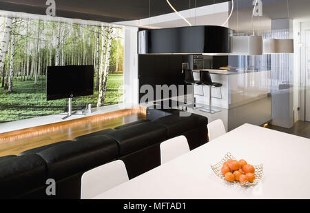 White dining table and chairs behind black leather sofa facing television in front of window Stock Photo