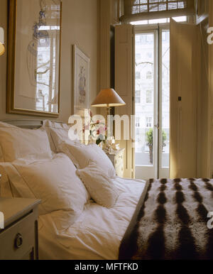 Pillows and striped fur throw on double bed in bedroom with lit bedside lamp and french doors with shutters. Stock Photo