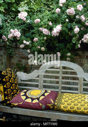 Wooden garden bench with cushions next to garden brick wall with climbing rose on wall fixed trellis Gardens detail seating rambling roses Stock Photo