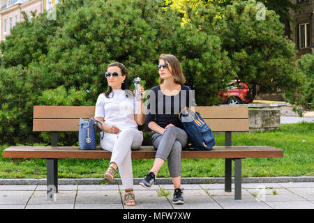 A girl and a young woman, wearing sunglasses, sit on a bench in the center of old Tallinn, and look in one direction. Stock Photo