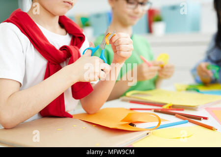 Kids sitting at school desk and cutting colorful paper with scissors while making origami on art lesson Stock Photo