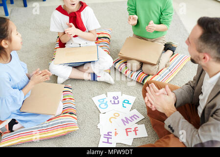 Primary school children and their male teacher sitting on the floor in classroom, playing games and learning alphabet letters Stock Photo