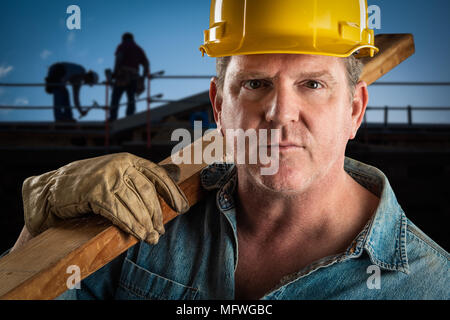 Serious Contractor in Hard Hat Carrying Wood Plank At Construction Site. Stock Photo