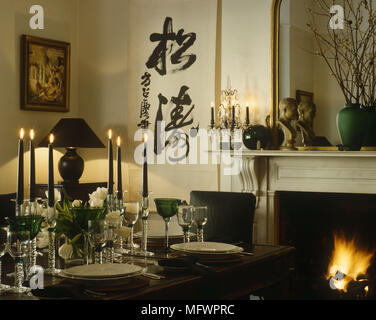 Wooden dining table with place setting next to fireplace with ornaments on mantelpiece and oriental characters painted on wall Stock Photo