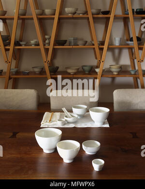 White oriental crockery placed on wooden dining table with upholstered chairs in front of wooden shelving unit Stock Photo