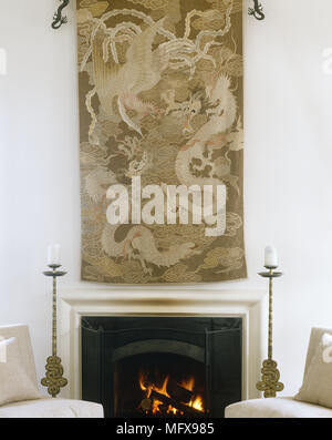 Oriental wall hanging above fireplace with lit fire with pair of candlesticks either side Stock Photo