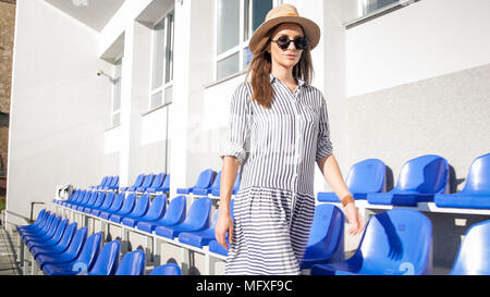 Beautiful stylish woman in summer hat walking between empty rows of seats Stock Photo