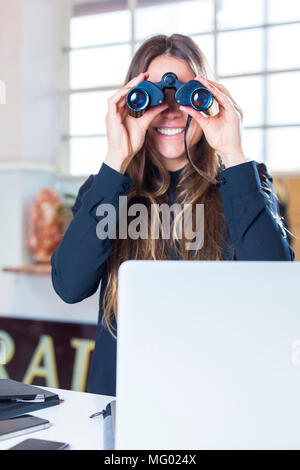 Deutschland, München, Portrait Geschäftsfrau im Büro vor Laptop durch Fernglas schauend Stock Photo