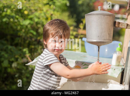 Cute boy, 4 years old, in striped T-shirt washes his hands under washstand outdoors in countryside Stock Photo