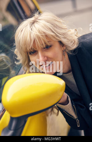 Young attractive blond woman paints her lips looking at the side mirror of a yellow car Stock Photo