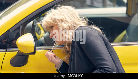 Young attractive blond woman paints her lips looking at the side mirror of a yellow car Stock Photo